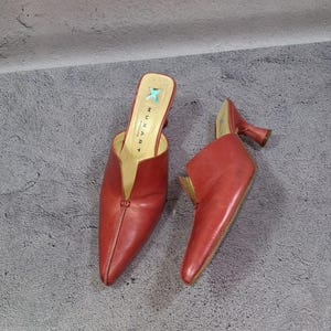 Peut inclure: Une paire de mules en cuir rouge avec un bout pointu et un talon bobine évasé. La semelle intérieure porte l'inscription MUXART BARCELONA avec un symbole de croix bleu clair. Les chaussures présentent une ouverture en V profond sur le dessus.