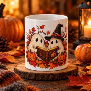 May include: A white ceramic mug featuring a graphic illustration of two ghosts reading an open book together. One ghost wears a black witch hat. The design includes orange pumpkins and vibrant red and orange autumn leaves surrounding them.
