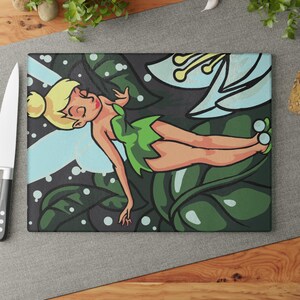 Könnte beinhalten: Ein rechteckiges Glasschneidebrett mit einer grafischen Illustration einer blonden Fee mit Dutt, die ein grünes Kleid trägt und zwischen großen dunkelgrünen Blättern sowie einer hellblauen Blume mit gelben Staubgefäßen vor dunklem Hintergrund ruht.