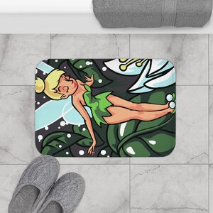 May include: A rectangular bath mat featuring a cartoon illustration of a blonde fairy with a green dress and translucent wings, resting among large dark green leaves and a white flower with yellow stamens against a dark grey background.