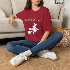 May include: A dark red short-sleeved t-shirt featuring the white text BASIC WITCH printed above a white graphic of a silhouette of a witch flying on a broomstick. The shirt is paired with blue denim jeans and white lace-up sneakers.