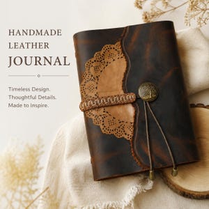 Könnte beinhalten: Ein dunkelbraunes Lederjournal mit einer beigen, spitzenartigen Ausschnittauflage, einem genähten Lederriemen und einem runden Messingknopf mit Baummotiv. Der Text lautet: HANDMADE LEATHER JOURNAL. Timeless Design. Thoughtful Details. Made to Inspire.