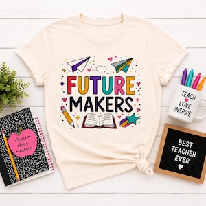 May include: A cream-colored t-shirt featuring a colorful graphic with the text "FUTURE MAKERS" in bold letters. The design includes illustrations of paper airplanes, a pencil, an open book, a star, and small hearts in various bright colors.