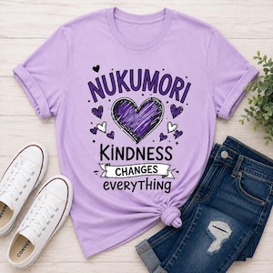 May include: A light purple short-sleeved t-shirt featuring a dark purple heart graphic in the center. The text reads NUKUMORI, KINDNESS CHANGES everything, surrounded by small heart icons. The shirt is displayed with white sneakers and blue jeans.