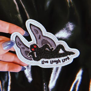 May include: An embroidered patch featuring a black silhouette of a creature with red eyes reclining in front of large, light purple moth wings. The patch has a white border and includes the black embroidered text "Live Laugh Lurk" at the bottom.
