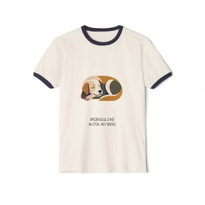 May include: A cream-colored ringer t-shirt with dark navy blue trim on the collar and sleeves. The center features a graphic of a sleeping dog in shades of brown, tan, and dark green, with the text "DEBUGGING WITH MY DOG" printed below it.
