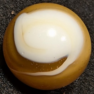 May include: A spherical glass marble featuring a translucent amber-colored base with an opaque white swirl pattern that curves across the surface. The glass has a smooth, polished texture with visible internal bubbles and light reflections.