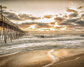 May include: A wooden pier extends out over a sandy beach with foamy waves crashing against the shore. The sky is a mix of gray and golden clouds with the sun peeking through.