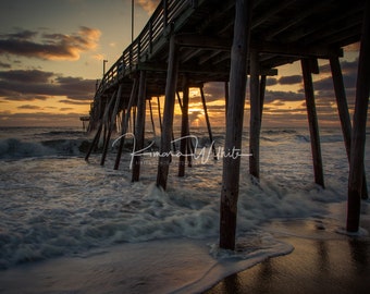 May include: A wooden pier extends over a sandy beach with foamy waves crashing against the supports. The sky is a vibrant orange and pink as the sun rises over the horizon.