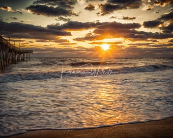 May include: A scenic photograph of a sunrise over the ocean. The sun, partially obscured by clouds, casts golden rays across the water. A wooden pier extends into the sea on the left. The sky is filled with dramatic clouds.
