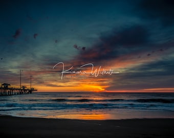 May include: A scenic view of a wooden pier extending into the ocean at sunset. The sky is a vibrant mix of orange, pink, and blue, with clouds reflecting the warm colors. The water is calm and reflects the sky's colors.