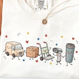 Può includere: Una t-shirt color crema con etichetta Comfort Colors in taglia S. La grafica presenta personaggi dei cartoni animati tra cui un furgone per le consegne, una scatola di cartone, un carrello, uno scanner di codici a barre e un bicchiere di caffè, circondati da piccole stelle e cuori.