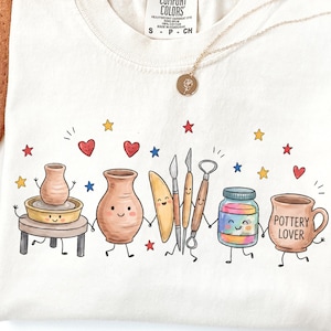May include: A cream-coloured t-shirt featuring a graphic illustration of anthropomorphic pottery tools and vessels holding hands. The design includes a pottery wheel, vases, a jar, and a mug with the text Pottery Lover, surrounded by colourful stars and hearts.