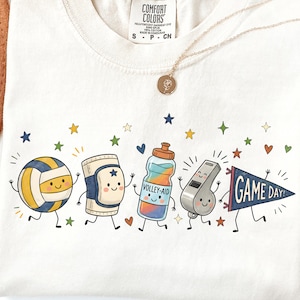 Peut inclure: Un t-shirt crème arborant un motif de cinq personnages de volley-ball dessinés : un ballon, une genouillère, une gourde marquée VOLLEY-AID, un sifflet et un fanion avec le texte GAME DAY, le tout entouré de petites étoiles et de cœurs.