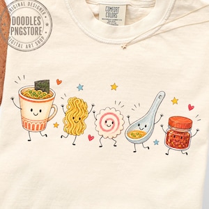 Könnte beinhalten: Ein cremefarbenes T-Shirt mit Rundhalsausschnitt und einem Grafikdruck von fünf anthropomorphen Lebensmitteln: ein Becher Ramen, ein Bündel Nudeln, ein Narutomaki-Fischkuchen, ein Suppenlöffel und ein Glas Chiliöl, alle mit lächelnden Gesichtern.
