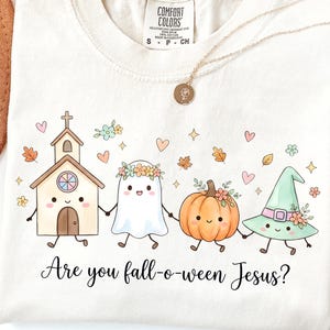 May include: A cream-colored t-shirt featuring a graphic of a cartoon church, a ghost with a flower crown, a pumpkin, and a witch hat holding hands. The text reads, Are you fall-o-ween Jesus? The shirt has a Comfort Colors tag showing size S.