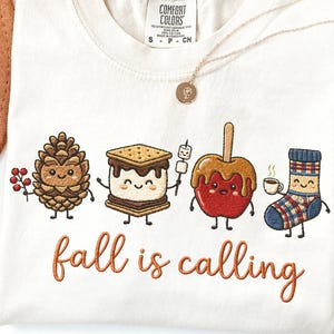 May include: A cream-colored t-shirt featuring an embroidered design of four anthropomorphic characters: a pinecone with berries, a s'more on a stick, a caramel apple, and a plaid sock holding a coffee cup, with the text "fall is calling" below.
