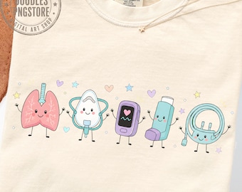May include: A cream-colored t-shirt featuring a graphic print of five cartoon medical items with smiling faces: pink lungs, a white oxygen mask, a purple pulse oximeter, a blue inhaler, and a light blue nasal cannula, accented by small stars and hearts.