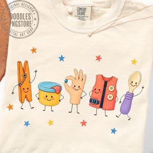 Puede incluir: Una camiseta de color crema con un gr&aacute;fico de cinco personajes antropom&oacute;rficos: una pinza de ropa naranja, un recipiente amarillo, una mano, un chaleco naranja y una cuchara morada. El dise&ntilde;o incluye peque&ntilde;as estrellas coloridas y el texto Comfort Colors.
