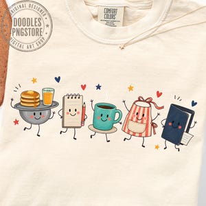 May include: A cream-colored t-shirt featuring a graphic print of five anthropomorphic characters: a bowl with pancakes and juice, a notepad with a pencil, a teal coffee mug, a red and white striped apron, and a dark blue book, all with faces.