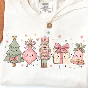 May include: A cream-colored t-shirt featuring a graphic print of five cartoon Christmas characters: a green tree, a pink ornament, a nutcracker, a gift box, and a bell, all with smiling faces, surrounded by small colorful stars and hearts.