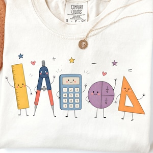May include: A cream-colored t-shirt with a graphic print of five anthropomorphic school supplies including a yellow ruler, a blue and red compass, a blue calculator, a purple pie chart, and an orange triangle, accented by small stars and hearts.