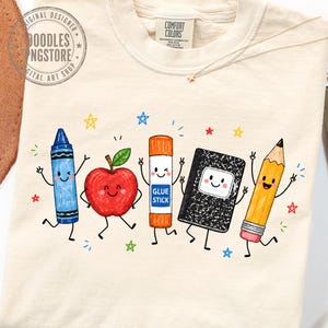May include: A cream-colored t-shirt featuring a graphic print of five anthropomorphic school supplies: a blue crayon, a red apple, an orange glue stick labeled GLUE STICK, a black and white composition notebook, and a yellow pencil with a pink eraser.