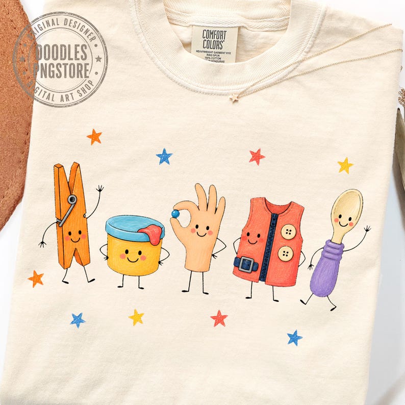 Puede incluir: Una camiseta de color crema con un gr&aacute;fico de cinco personajes antropom&oacute;rficos: una pinza de ropa naranja, un recipiente amarillo, una mano, un chaleco naranja y una cuchara morada. El dise&ntilde;o incluye peque&ntilde;as estrellas coloridas y el texto Comfort Colors.
