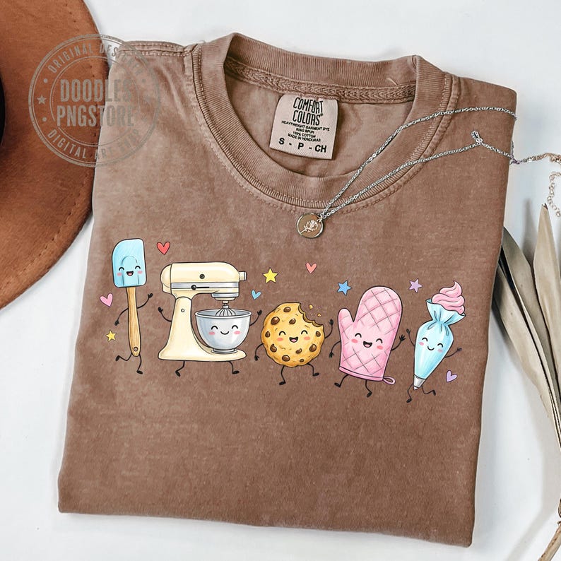 May include: A brown short-sleeve t-shirt featuring a graphic print of five animated baking items: a blue spatula, a cream stand mixer, a chocolate chip cookie, a pink oven mitt, and a blue piping bag, all with smiling faces and limbs.