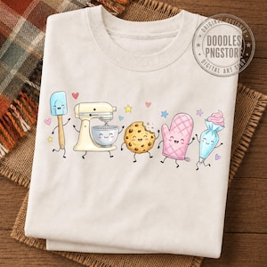 May include: A cream-colored t-shirt featuring a graphic print of anthropomorphic baking items including a spatula, a stand mixer, a chocolate chip cookie, an oven mitt, and a piping bag, all decorated with small colorful stars and hearts.