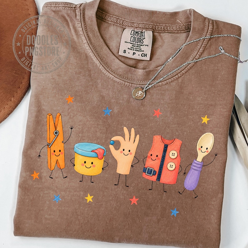 Puede incluir: Una camiseta marr&oacute;n de manga corta con un estampado gr&aacute;fico de cinco personajes de dibujos animados: una pinza de ropa naranja, un recipiente amarillo, una mano haciendo un gesto de ok, un chaleco naranja y una cuchara morada, rodeados de estrellas.