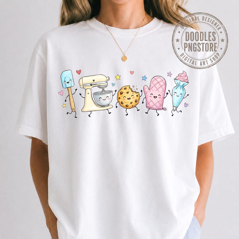 May include: A white short-sleeved t-shirt featuring a graphic print of animated baking tools including a spatula, a stand mixer, a chocolate chip cookie, an oven mitt, and a piping bag, all with smiling faces, surrounded by small colorful stars and hearts.