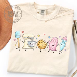 May include: A cream-colored t-shirt featuring a graphic print of animated baking tools including a spatula, a stand mixer, a chocolate chip cookie, an oven mitt, and a piping bag, all with smiling faces, surrounded by small colorful stars and hearts.