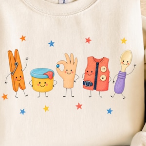 Puede incluir: Una sudadera de color crema con un estampado gr&aacute;fico de cinco personajes antropom&oacute;rficos: una pinza de ropa naranja, un recipiente amarillo, una mano sosteniendo una canica azul, un chaleco rojo con botones y una cuchara morada, rodeados de estrellas.
