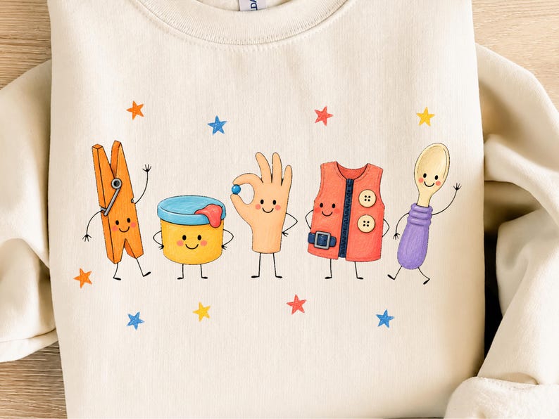 Puede incluir: Una sudadera de color crema con un estampado gr&aacute;fico de cinco personajes antropom&oacute;rficos: una pinza de ropa naranja, un recipiente amarillo, una mano sosteniendo una canica azul, un chaleco rojo con botones y una cuchara morada, rodeados de estrellas.
