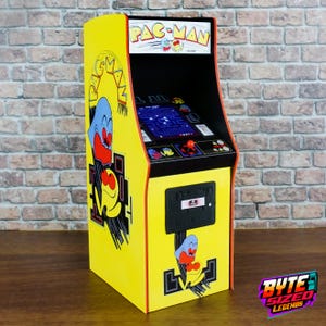 Può includere: Un cabinato arcade Pac-Man giallo con grafiche del personaggio iconico e fantasmi sui pannelli laterali e frontali. L'unità include un'insegna superiore con il logo Pac-Man, un piccolo schermo che mostra il gioco, un joystick rosso e dei pulsanti.