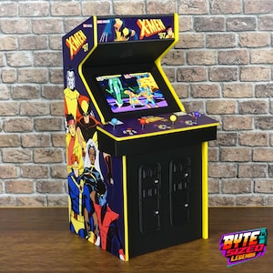Könnte beinhalten: Ein Miniatur-Spielautomat X-Men '97 mit einem Bildschirm, der ein Videospiel anzeigt, einem Bedienfeld mit drei Joysticks und bunten Knöpfen sowie seitlichen Grafiken, die verschiedene X-Men-Charaktere wie Wolverine und Storm darstellen.