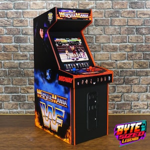 Può includere: Un cabinato arcade in miniatura con marchio WrestleMania, uno schermo che mostra un gioco di wrestling, un pannello di controllo con un joystick blu e uno rosso, e grafiche laterali raffiguranti un wrestler. Include i loghi Midway e WWF.
