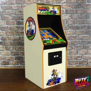 May include: A miniature Rally-X arcade cabinet featuring a cream-colored exterior with colorful graphics of a race car. The unit includes a top marquee, a screen displaying a blue and orange maze, a red joystick, and a black coin door on the front.