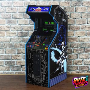 Puede incluir: Una máquina de arcade en miniatura de Star Wars con exterior negro, detalles en azul y gráficos detallados del espacio. La marquesina muestra el logotipo de Atari Star Wars, mientras que los paneles laterales presentan a Darth Vader y naves X-wing.