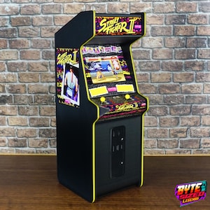 Puede incluir: Una máquina recreativa en miniatura de color negro con la marca Street Fighter II, bordes amarillos y una pantalla que muestra el juego. El panel lateral muestra una ilustración de un personaje y el panel de control incluye dos joysticks.