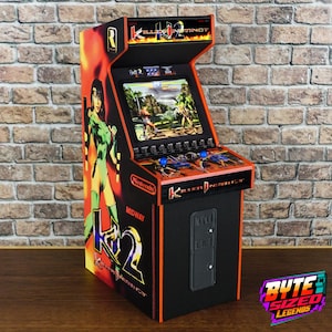 Könnte beinhalten: Ein Tisch-Arcade-Automat mit Killer Instinct 2-Branding und einem Bildschirm, der ein Kampfspiel anzeigt. Die Seitenwand zeigt eine Charakterillustration, während das Bedienfeld zwei blaue Joysticks und Tasten enthält. Das Gerät ist schwarz mit orangefarbenen Akzenten sowie Nintendo- und Midway-Logos.