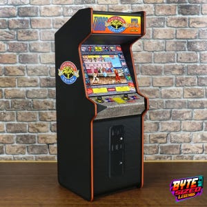 Puede incluir: Una máquina arcade de sobremesa negra con la marca Street Fighter II Champion Edition. La marquesina muestra el título del juego, mientras que la pantalla muestra una escena de lucha. El panel de control incluye dos joysticks y botones naranjas.