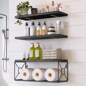 May include: Three black wall-mounted shelves arranged vertically. The top two are solid planks with metal brackets, holding a potted plant, bottles, jars, and tubes. The bottom shelf features a black metal wire frame holding three rolls of toilet paper.