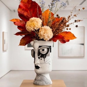 May include: A white ceramic vase shaped like a human face with black line art details, including a prominent eye, dark lips, and swirling patterns at the base. The vase is filled with a vibrant arrangement of large red leaves and beige flowers.