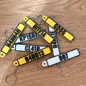 May include: A collection of rectangular keychains shaped like license plates in yellow and white with black borders. The keychains feature raised black lettering including H4NN4H, 0I1V1A, GDOG, KP15RE, H4KL1E, CL41R, B4DD13, and 007, each attached to a metal split ring.