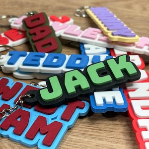 May include: A collection of multi-coloured 3D printed plastic keychains featuring raised block lettering. The keychains display various names and words including JACK, DAD, TEDDY, and NANNY in contrasting colour combinations with metal key rings.