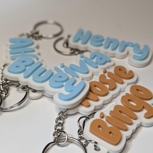 May include: A collection of custom keychains featuring raised lettering on a white base. The names Henry, Olivia, Rosie, and Bingo are displayed in light blue or tan 3D-style text. Each keychain includes a silver metal split ring and chain.