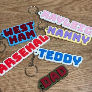 May include: A collection of six custom keychains featuring raised block lettering in various colors. The keychains display the words West Ham, Arsenal, Kayleigh, Nanny, Teddy, and Dad, each attached to a silver metal split ring for keys.