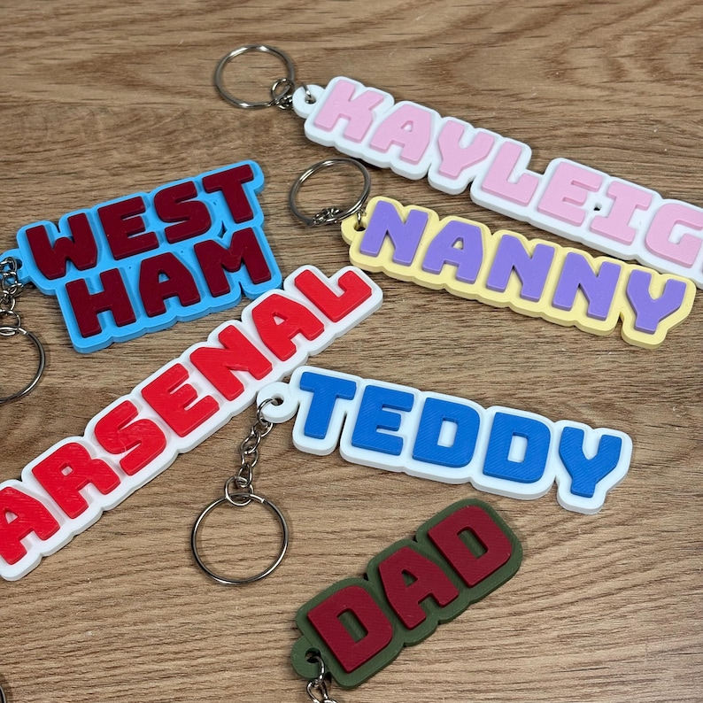May include: A collection of six custom keychains featuring raised block lettering in various colors. The keychains display the words West Ham, Arsenal, Kayleigh, Nanny, Teddy, and Dad, each attached to a silver metal split ring for keys.